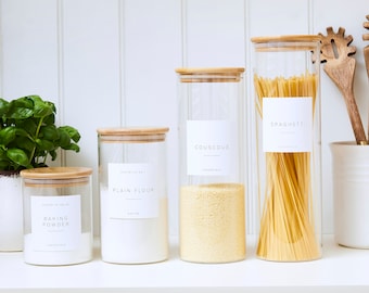 May include: Four glass jars with bamboo lids and white labels. The labels have the words "Baking Powder", "Plain Flour", "Couscous", and "Spaghetti" written on them. The jars are sitting on a white countertop.