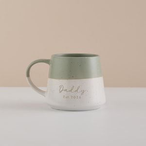 May include: A two-toned ceramic mug with a sage green top and white speckled bottom. The mug has a curved handle and the word "Daddy" is written in a script font, with "Est. 2026" below it.