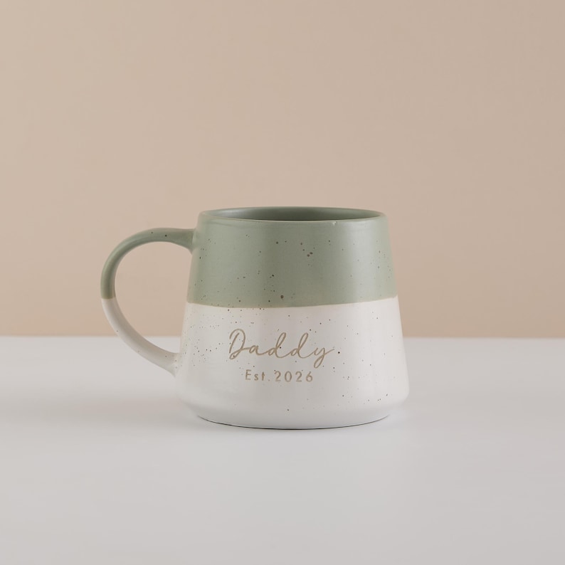 May include: A two-toned ceramic mug with a sage green top and white speckled bottom. The mug has a curved handle and the word "Daddy" is written in a script font, with "Est. 2026" below it.