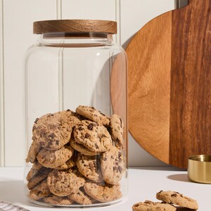 Peut inclure: Un bocal en verre transparent avec un couvercle en bois rempli de biscuits aux p&eacute;pites de chocolat. Le bocal est pos&eacute; sur une surface blanche avec un tissu &agrave; carreaux brun et blanc.