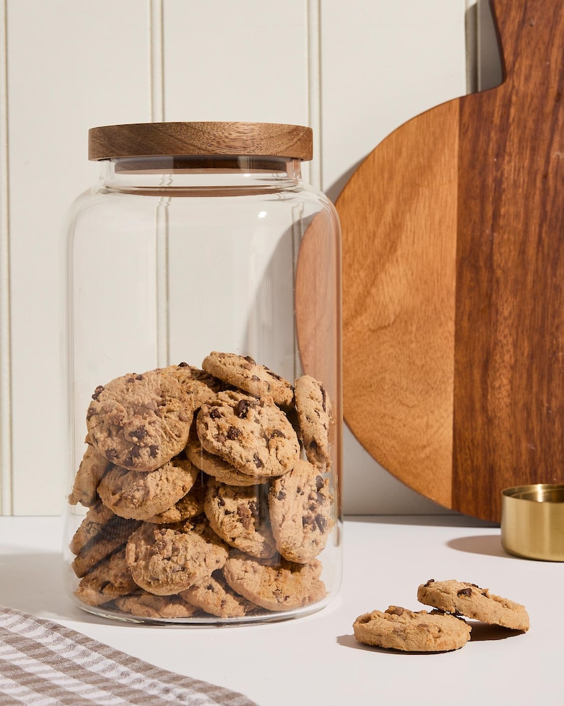 Peut inclure: Un bocal en verre transparent avec un couvercle en bois rempli de biscuits aux p&eacute;pites de chocolat. Le bocal est pos&eacute; sur une surface blanche avec un tissu &agrave; carreaux brun et blanc.