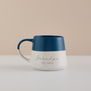 May include: A ceramic mug with a navy blue top and a speckled white bottom. The word "Grandpa" is written in a script font, with "Est. 2026" below it. The handle is white with a navy blue accent.