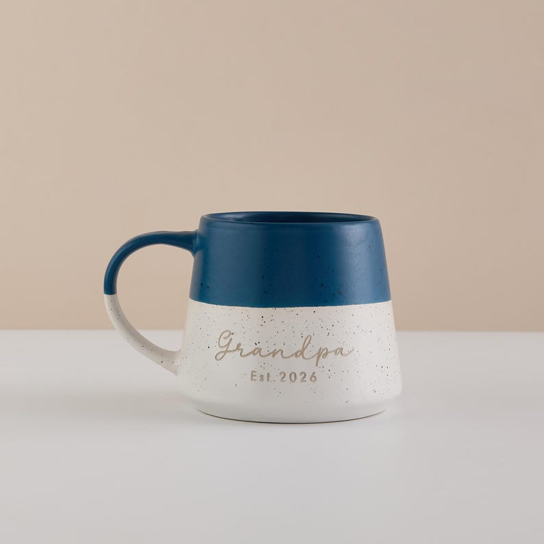 May include: A ceramic mug with a navy blue top and a speckled white bottom. The word "Grandpa" is written in a script font, with "Est. 2026" below it. The handle is white with a navy blue accent.