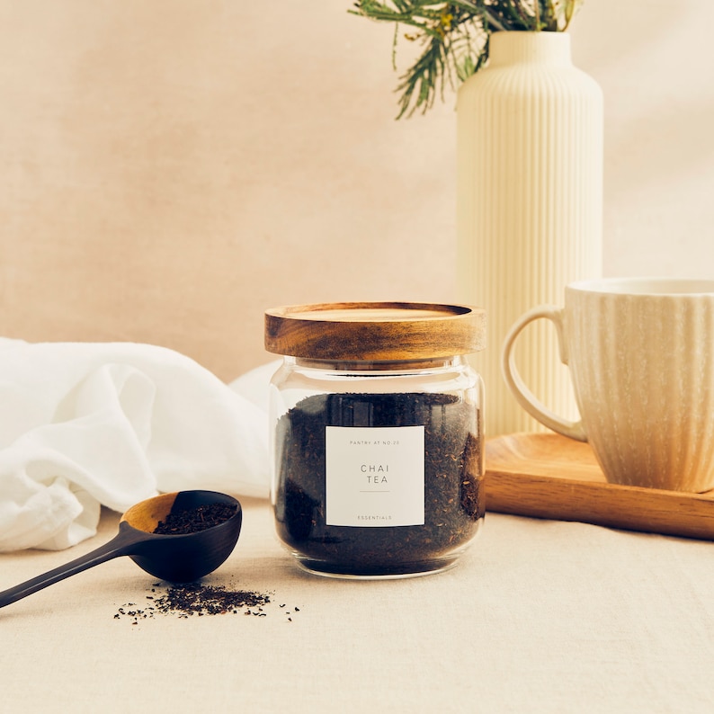 May include: A glass jar with a wooden lid filled with loose leaf chai tea. The jar has a label that reads "Chai Tea" and is sitting on a light brown surface. There is a wooden spoon with tea leaves in it and a white ceramic mug in the background.
