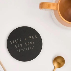 May include: A round, black coaster with the text "BELLE & MAX NEW HOME 12/03/2024" in a light color. A gold spoon and a brown mug with a tan rim are also visible.