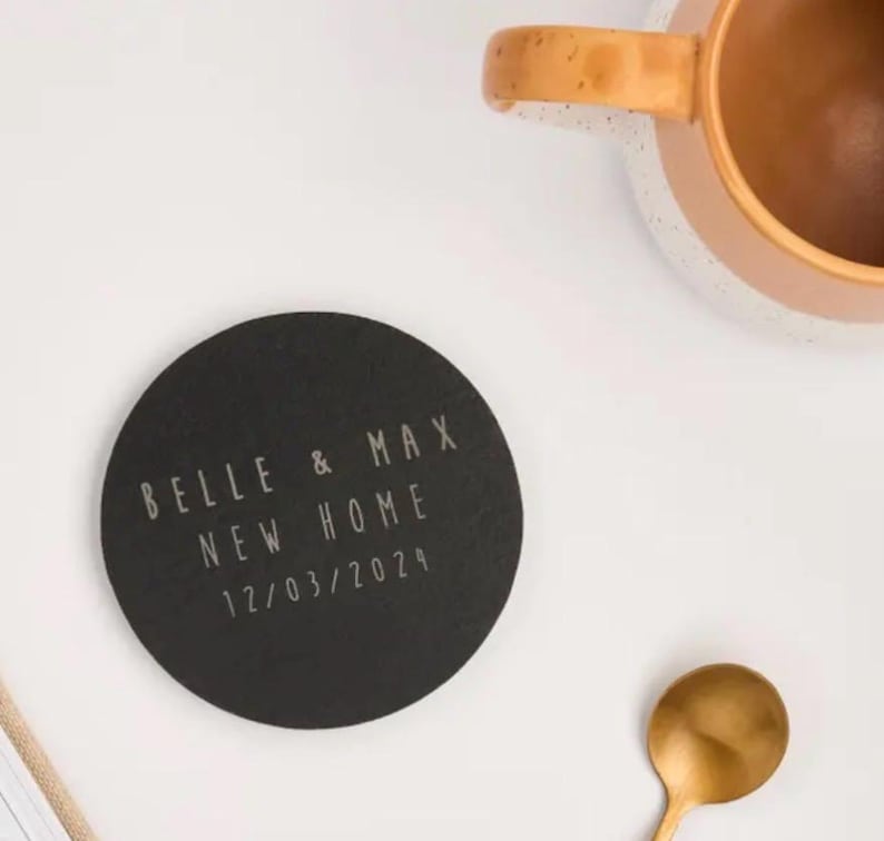 May include: A round, black coaster with the text "BELLE & MAX NEW HOME 12/03/2024" in a light color. A gold spoon and a brown mug with a tan rim are also visible.