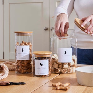 May include: Three glass jars with bamboo lids filled with dog treats. The jars are labelled "Dani's Treats", "Luna's Treats", and "Bella's Food". Each label features a silhouette of a dog or cat. The jars are on a wooden table with a few dog treats scattered around.