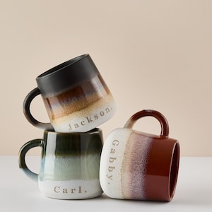 May include: Three ceramic mugs with a gradient glaze in black, green, and red. The mugs are stacked on top of each other. The mugs are personalized with the names "Jackson", "Gabby", and "Carl".