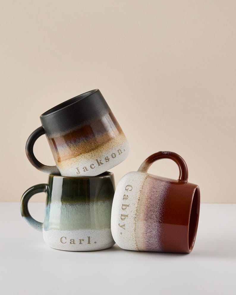 May include: Three ceramic mugs with a gradient glaze in black, green, and red. The mugs are stacked on top of each other. The mugs are personalized with the names "Jackson", "Gabby", and "Carl".