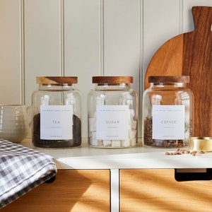 May include: Three glass jars with wooden lids and labels that read "Tea", "Sugar", and "Coffee". The jars are filled with tea, sugar cubes, and coffee beans, respectively. The jars are sitting on a white countertop.