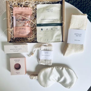 May include: A gift box filled with self-care items. Includes bath salts, a face mask, wool socks, a soap bag, a body bar, scented tea lights, and an eye mask. The products are branded with the name "Arthur Betsy". The box is filled with straw.