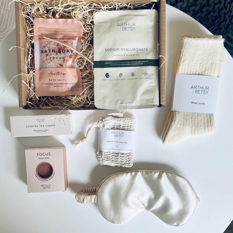 May include: A gift box filled with self-care items. Includes bath salts, a face mask, wool socks, a soap bag, a body bar, scented tea lights, and an eye mask. The products are branded with the name "Arthur Betsy". The box is filled with straw.