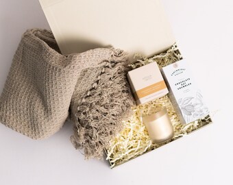 Puede incluir: Una caja de regalo llena de una manta de lana beige, una vela y una caja de migas de avena con chocolate. La caja está forrada con papel blanco triturado.