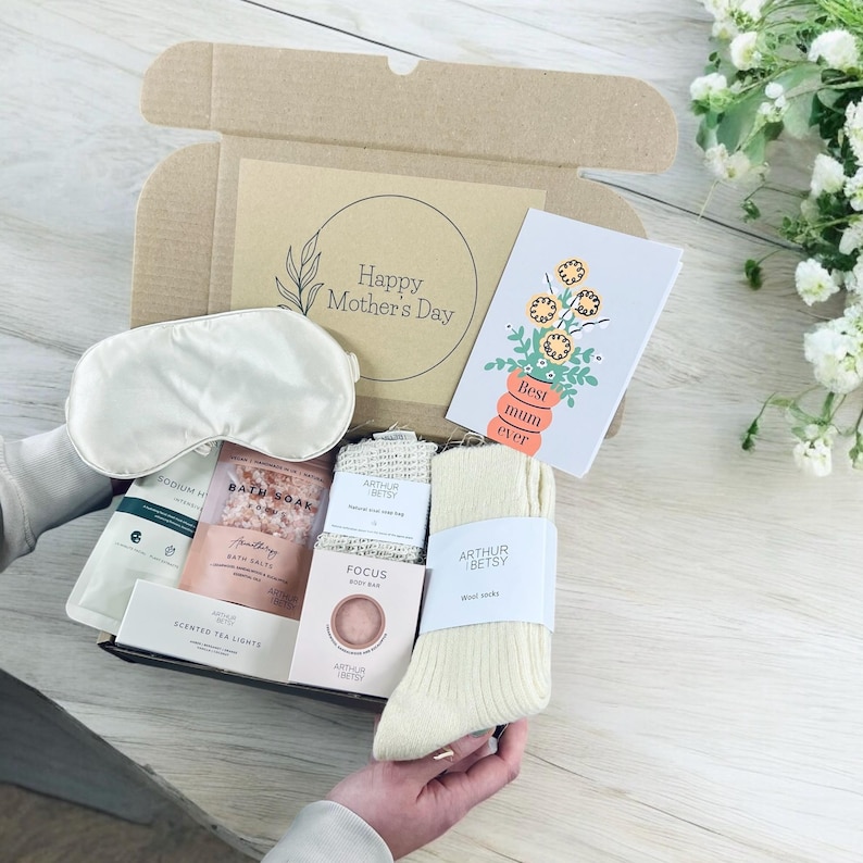 May include: A gift box with a variety of self-care items, including a sleep mask, bath salts, soap, socks, and scented tea lights. A card with the text "Best mum ever" is also included. The box is open, revealing the contents.
