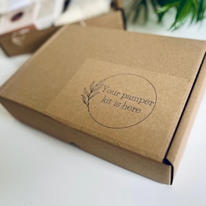 May include: A brown cardboard box with a sticker that reads "Your pamper kit is here" in a circular design. The box is closed and appears to be ready for shipping. Other boxes are visible in the background.