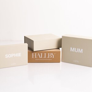 Pu&ograve; includere: Tre scatole regalo beige con scritte bianche. La scatola superiore dice "MUM", la scatola centrale dice "HALLBY" e la scatola inferiore dice "SOPHIE".