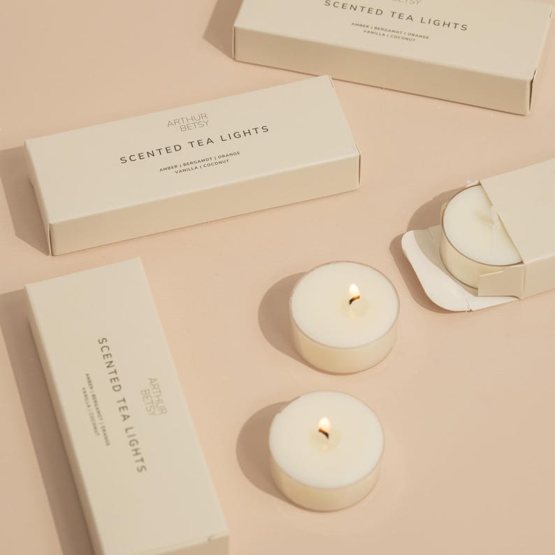 May include: Cream-colored tea light candles with lit flames, alongside rectangular boxes. The boxes are labeled "SCENTED TEA LIGHTS" with scent details. The brand name "ARTHUR BETSY" is also visible. The background is a soft, neutral color.