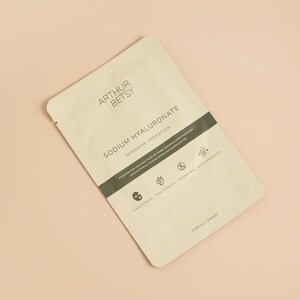 May include: A beige packet of Arthur & Betsy Sodium Hyaluronate intensive hydration facial sheet mask. The packet has a dark green band with text and icons. The bottom right corner reads 0.8 fl oz / 25ml. The background is a light peach color.