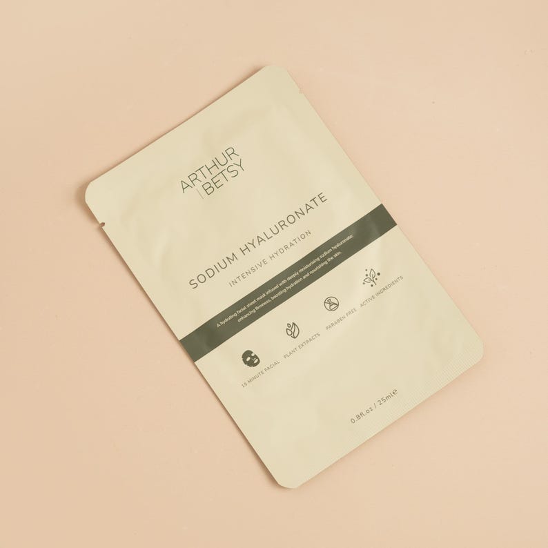 May include: A beige packet of Arthur & Betsy Sodium Hyaluronate intensive hydration facial sheet mask. The packet has a dark green band with text and icons. The bottom right corner reads 0.8 fl oz / 25ml. The background is a light peach color.