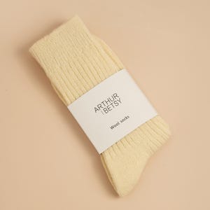 May include: A pair of pale yellow wool socks with ribbed texture, wrapped with a white label. The label features the text "ARTHUR | BETSY" above "Wool socks". The socks are folded and angled on a light beige surface.