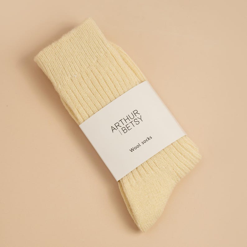 May include: A pair of pale yellow wool socks with ribbed texture, wrapped with a white label. The label features the text "ARTHUR | BETSY" above "Wool socks". The socks are folded and angled on a light beige surface.
