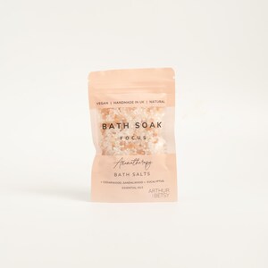 May include: A peach-colored pouch of bath salts with the text "BATH SOAK FOCUS Aromatherapy BATH SALTS". The pouch contains pink and white bath salts. The text "VEGAN | HANDMADE IN UK | NATURAL" is at the top.