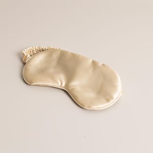 May include: A gold-colored sleep mask made of a smooth, satin-like material. The mask has a curved shape to fit around the eyes and a gathered elastic band. The sleep mask is designed to block out light for better sleep.