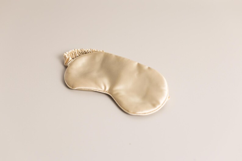 May include: A gold-colored sleep mask made of a smooth, satin-like material. The mask has a curved shape to fit around the eyes and a gathered elastic band. The sleep mask is designed to block out light for better sleep.