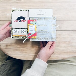 May include: A gift box filled with self-care items, including a cat-themed coaster that says "Cat Mum", two small bottles of tea, a bar of chocolate, and a self-heating eye mask with the words "Sensory Retreats" and "Divine Eyes".