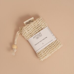 May include: A natural sisal soap bag with a drawstring closure and a wooden bead. The bag is made of woven beige fibers and features a white label with the text "ARTHUR | BETSY" and "Natural sisal soap bag."