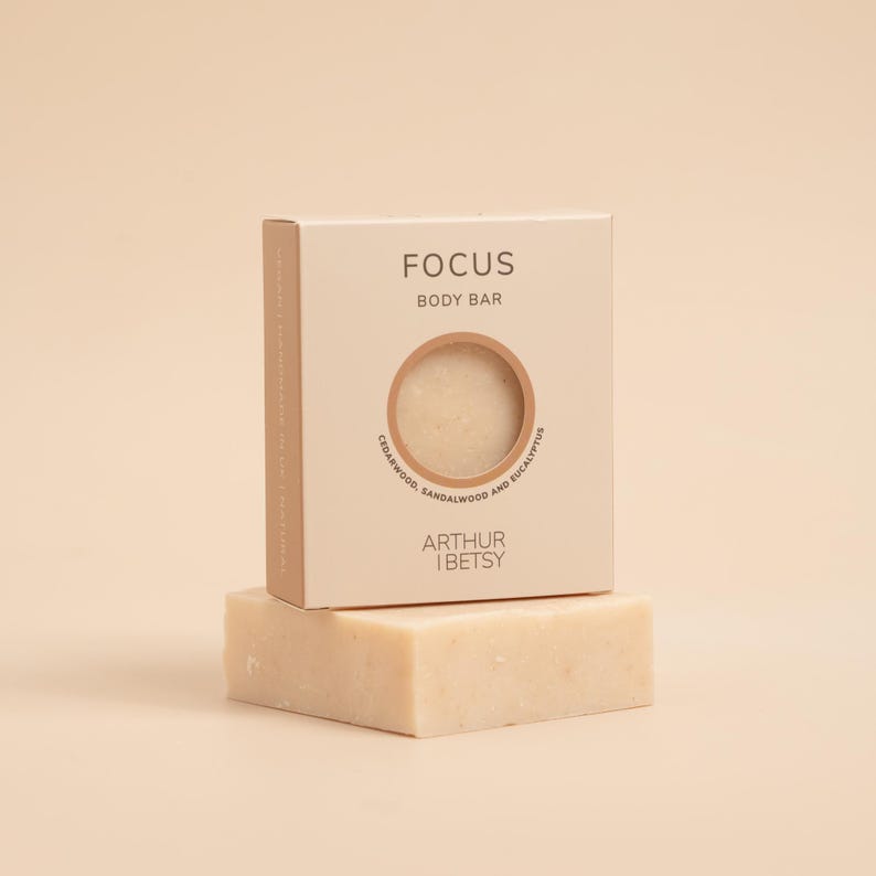 May include: A light beige body bar and its box. The box is labeled "FOCUS BODY BAR" with a circular window revealing the soap. The box also says "CEDARWOOD, SANDALWOOD AND EUCALYPTUS" and "ARTHUR & BETSY". The soap is rectangular.