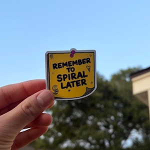 May include: A square yellow sticker shaped like a sticky note with a pink pushpin at the top. The text REMEMBER TO SPIRAL LATER is printed in black capital letters. The design includes small black hand-drawn doodles of a spiral and a heart.