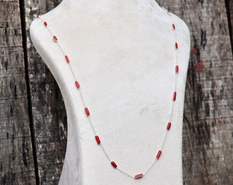 May include: A red and silver necklace with rectangular red stones on a white mannequin bust. The bust is on a wooden shelf.