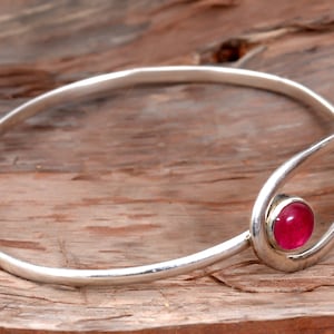 May include: A silver bangle bracelet with a single pink gemstone set in the center. The bracelet is open and has a simple, elegant design.