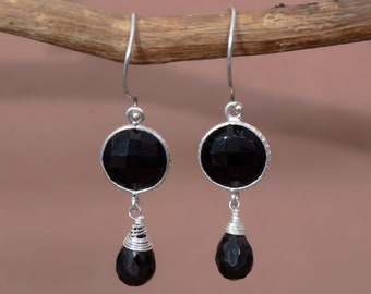 May include: A pair of silver dangle earrings with black faceted gemstone drops. The earrings feature a round, black gemstone with a silver bezel setting and a smaller, teardrop-shaped black gemstone hanging below.
