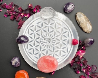 May include: A white selenite charging plate with a flower of life design and a crystal eye symbol. The plate is surrounded by various crystals, including pink, orange, purple, and brown.