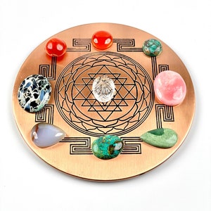 Könnte beinhalten: Eine Kupferplatte mit einem Sri Yantra-Design und acht Edelsteinen, die um einen klaren Kristall in der Mitte angeordnet sind. Die Edelsteine sind rot, rosa, grün, grau und weiß.