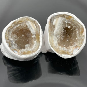 Puede incluir: Dos mitades de una geoda con un exterior blanco y un interior claro lleno de cristales. La geoda está envuelta en papel de seda blanco.