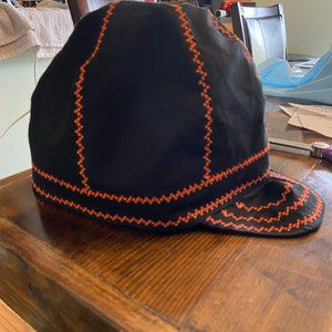 May include: Black welding cap with orange zigzag stitching. The cap has a rounded crown and a short, curved brim. The stitching outlines the seams and edges of the cap, creating a contrasting design. The cap is sitting on a wooden surface.