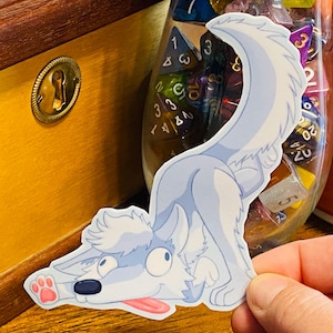 May include: A playful sticker featuring a light blue cartoon wolf in a playful pose, with its tongue sticking out. The wolf has a white fluffy tail and paws. The sticker is held in a hand, with a wooden box and dice in the background.