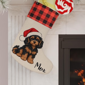 Peut inclure: Une chaussette de Noël beige avec un revers à carreaux rouges et noirs. La chaussette présente une illustration d'un chiot noir et feu portant un bonnet de Père Noël rouge, avec le prénom Max écrit en écriture cursive noire sous le chiot.