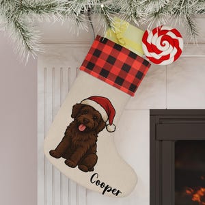 May include: A Christmas stocking with a red and black buffalo plaid cuff. The stocking features a brown dog wearing a Santa hat, with the name "Cooper" printed below. A red and white swirled lollipop is attached.