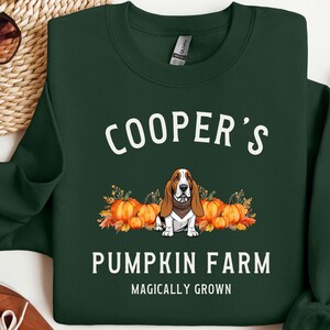 Könnte beinhalten: Dunkelgrünes Sweatshirt mit dem Text "COOPER'S PUMPKIN FARM" und einer Grafik eines Basset Hounds vor Kürbissen. Die Worte "MAGICALY GROWN" stehen unter dem Namen der Farm.