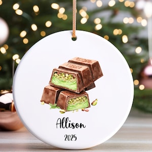 May include: White ceramic Christmas ornament with a watercolor illustration of chocolate bars with pistachio filling. The ornament has a gold string for hanging and features the name "Allison" and the year "2025".
