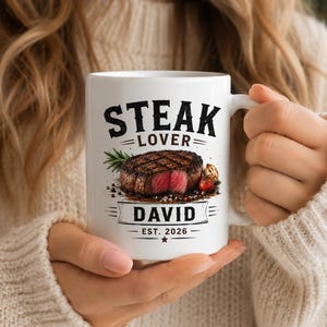 Op de afbeelding: Een witte keramische mok met een afbeelding van een gebakken biefstuk, een takje rozemarijn en een kleine tomaat. De tekst luidt STEAK LOVER, DAVID en EST. 2026 in zwarte letters. De mok is voorzien van een handvat aan de rechterkant.