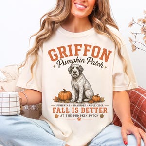 以下が含まれることがあります： グリフォンの犬、カボチャ、そして「Griffon Pumpkin Patch」の文字が描かれたクリーム色のTシャツ。デザインには「Fall is Better at the Pumpkin Patch」というフレーズが含まれています。
