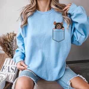 May include: Light blue sweatshirt featuring a graphic of a brown rat peeking out of a pocket. The pocket is outlined in black stitching. The sweatshirt is a solid light blue color.