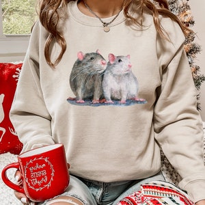 May include: A beige sweatshirt with a watercolor illustration of two rats. The rats are sitting next to each other and looking up. The sweatshirt has a crew neck and long sleeves.