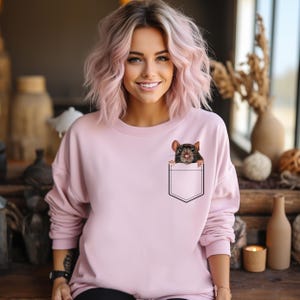 May include: A light pink sweatshirt featuring a graphic of a rat peeking out of a pocket. The rat is brown and black with a detailed face. The sweatshirt has long sleeves and a crew neck.