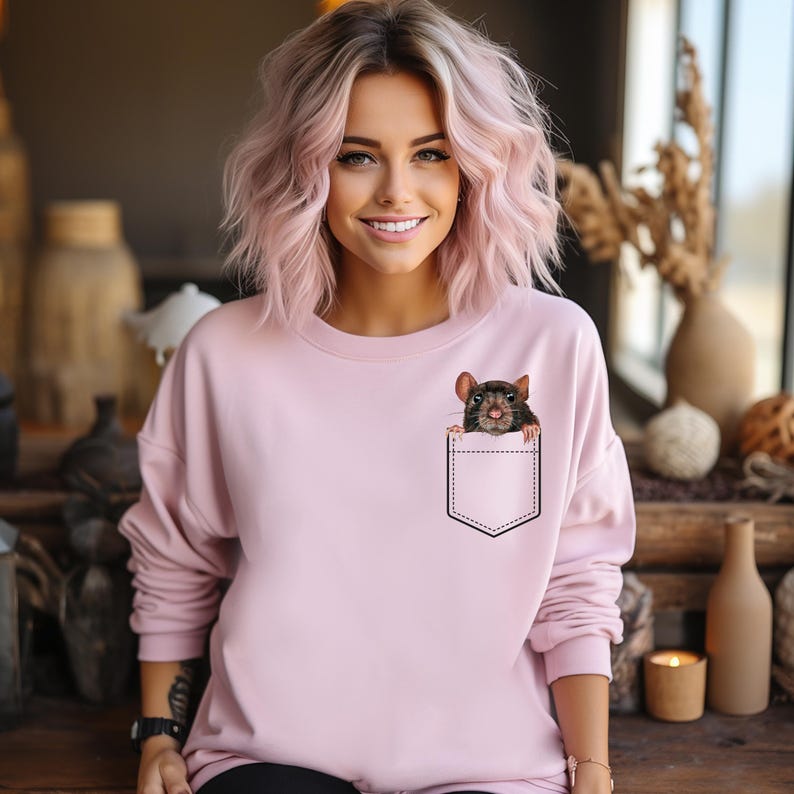 May include: A light pink sweatshirt featuring a graphic of a rat peeking out of a pocket. The rat is brown and black with a detailed face. The sweatshirt has long sleeves and a crew neck.